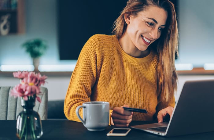 A person smiling while sitting at a table in front of a computer and holding a payment card.